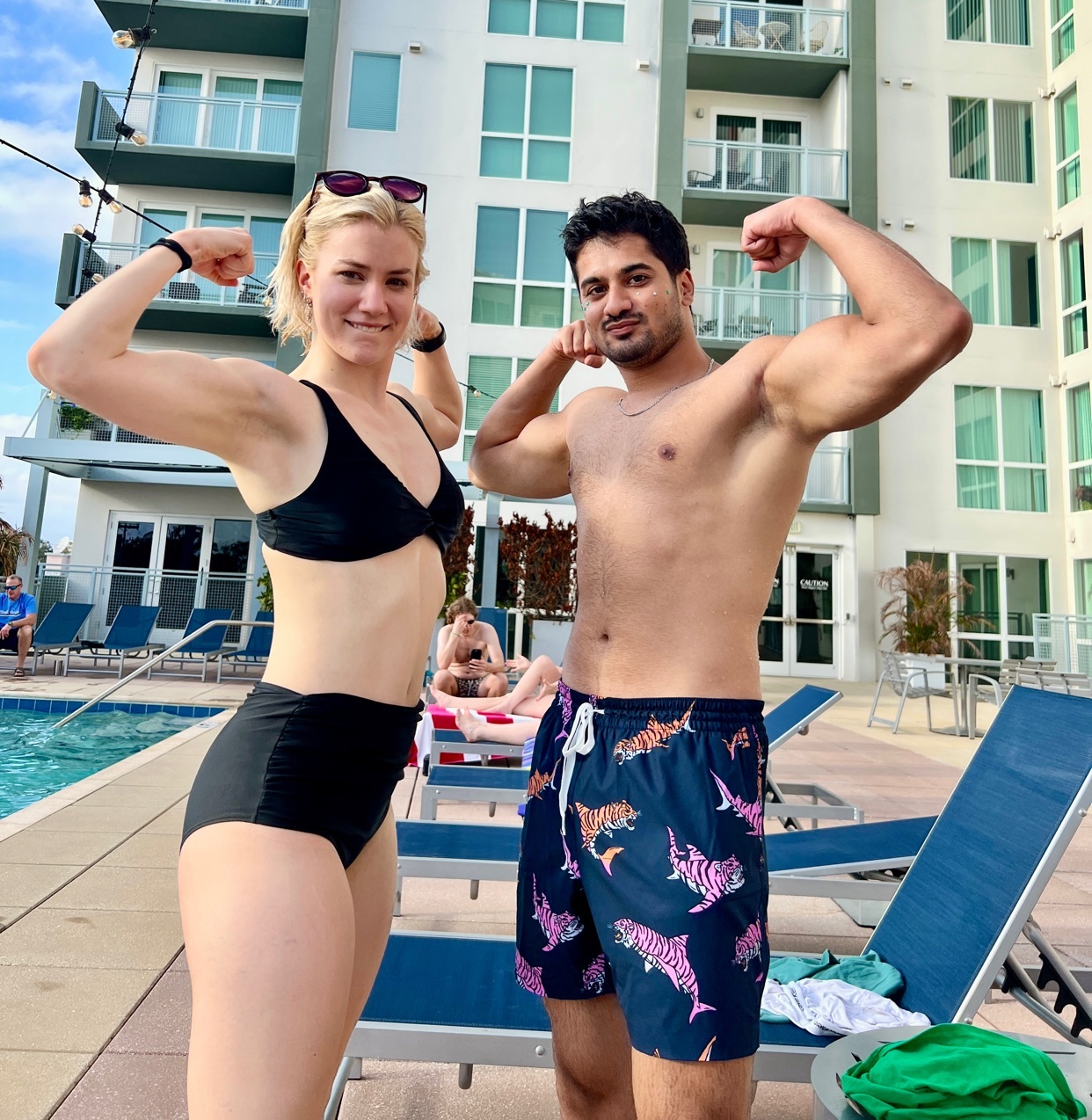 A muscular couple poses by a pool, flexing their biceps. The woman wears a black bikini, and the man wears patterned swim trunks.