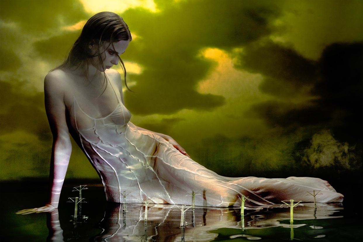 A surreal digital art piece featuring a woman in a flowing dress, partially submerged in water. The artwork has a dreamlike quality with a dark, cloudy background.