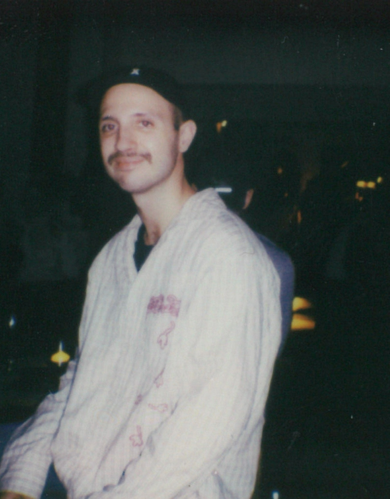A man poses for a photo, wearing a cap and a patterned shirt. The photo is taken in a vintage style.