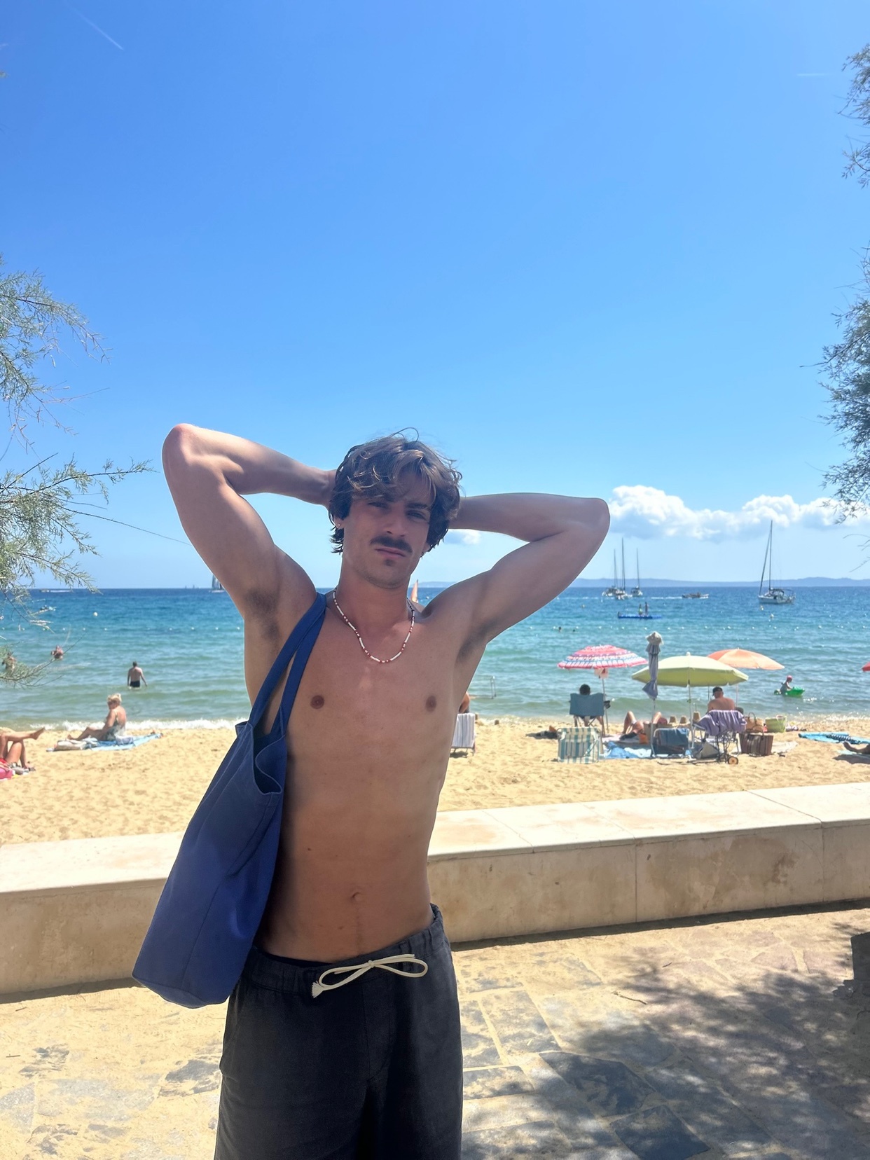 A man poses shirtless on a sunny beach. The photo captures a summer vacation vibe with the ocean and sky in the background.