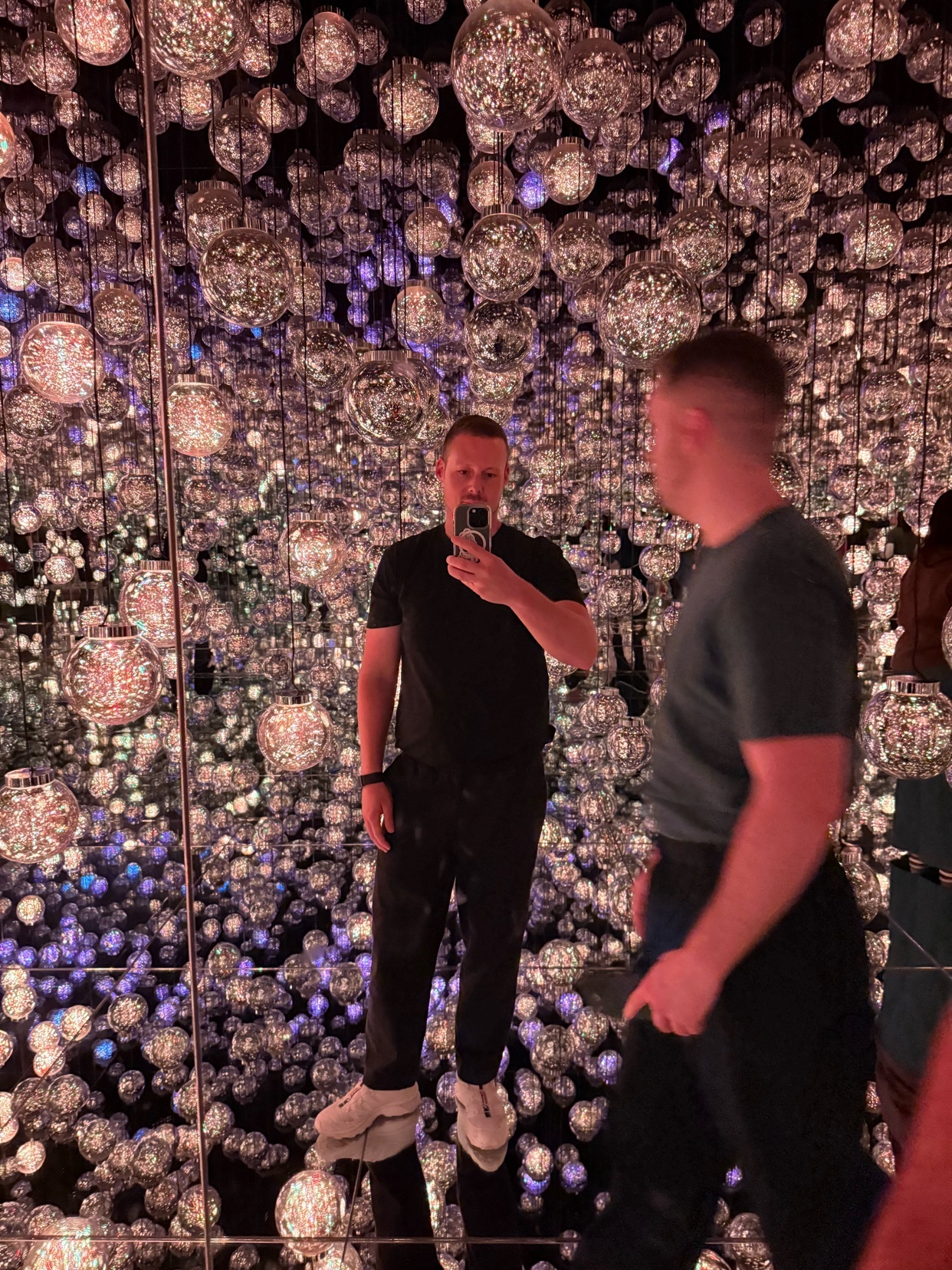 A man takes a mirror selfie in an art installation filled with reflective disco balls and purple lights.