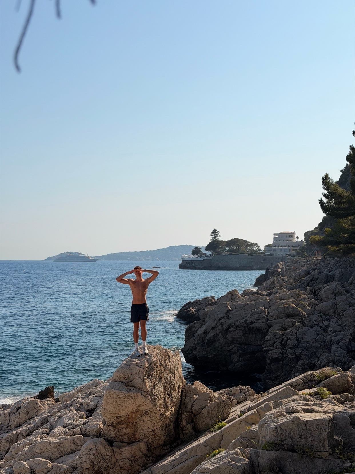 A shirtless man stands on a rock overlooking the ocean, with his hands behind his head. The rocky coastline and clear blue sky suggest a summer day.