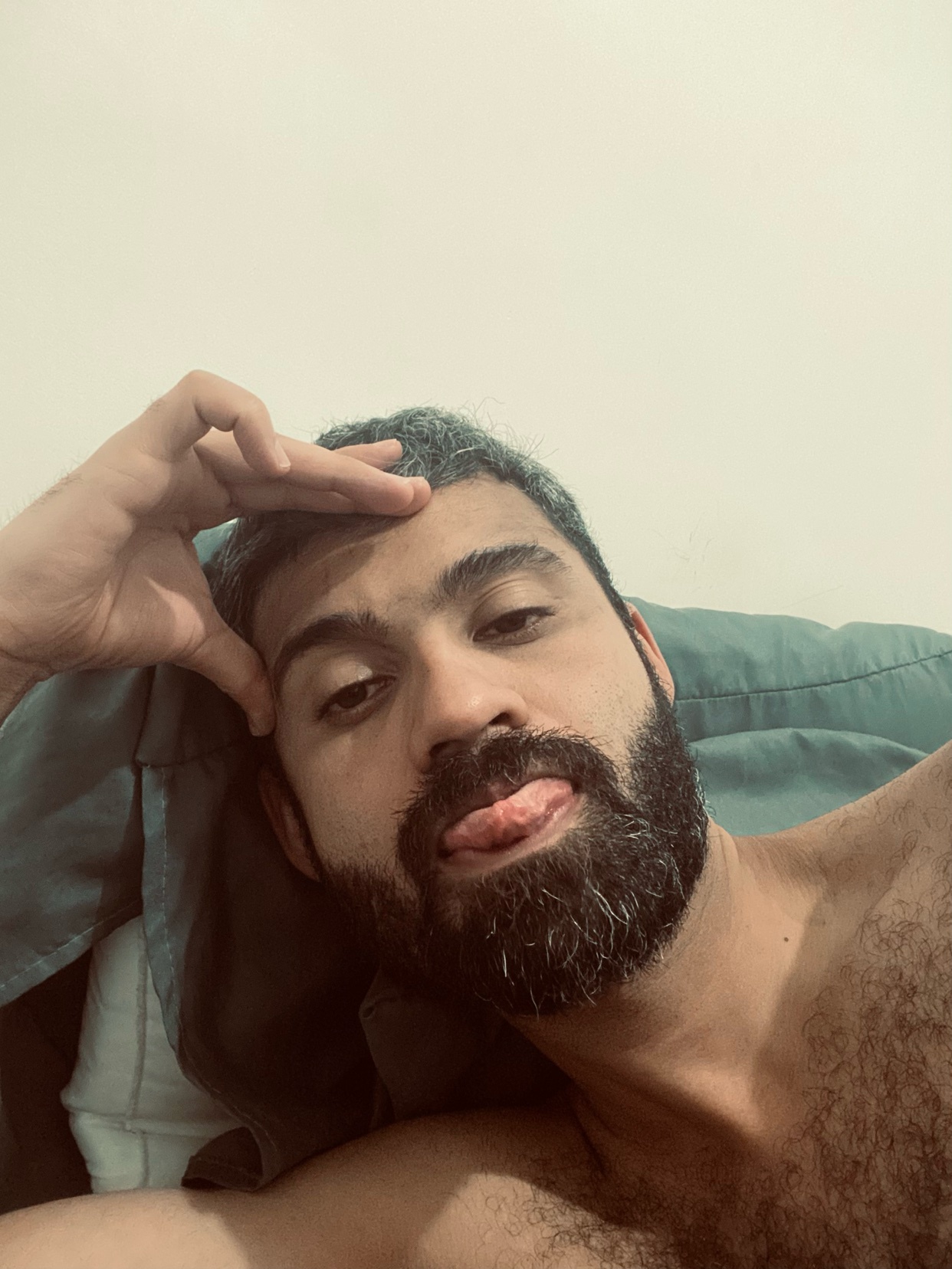 A man is taking a selfie, sticking his tongue out. He is shirtless and has a beard.