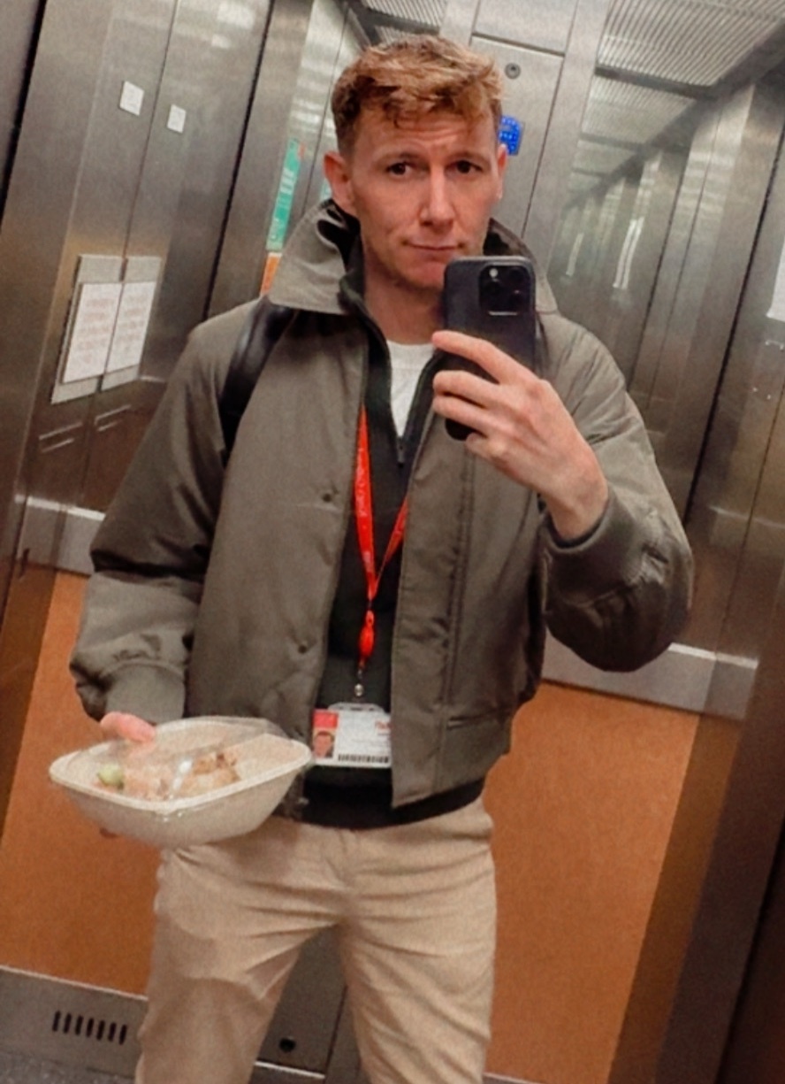 A man takes a selfie in an elevator while holding a takeout container of food. He is wearing a bomber jacket over a white t-shirt and khaki pants.