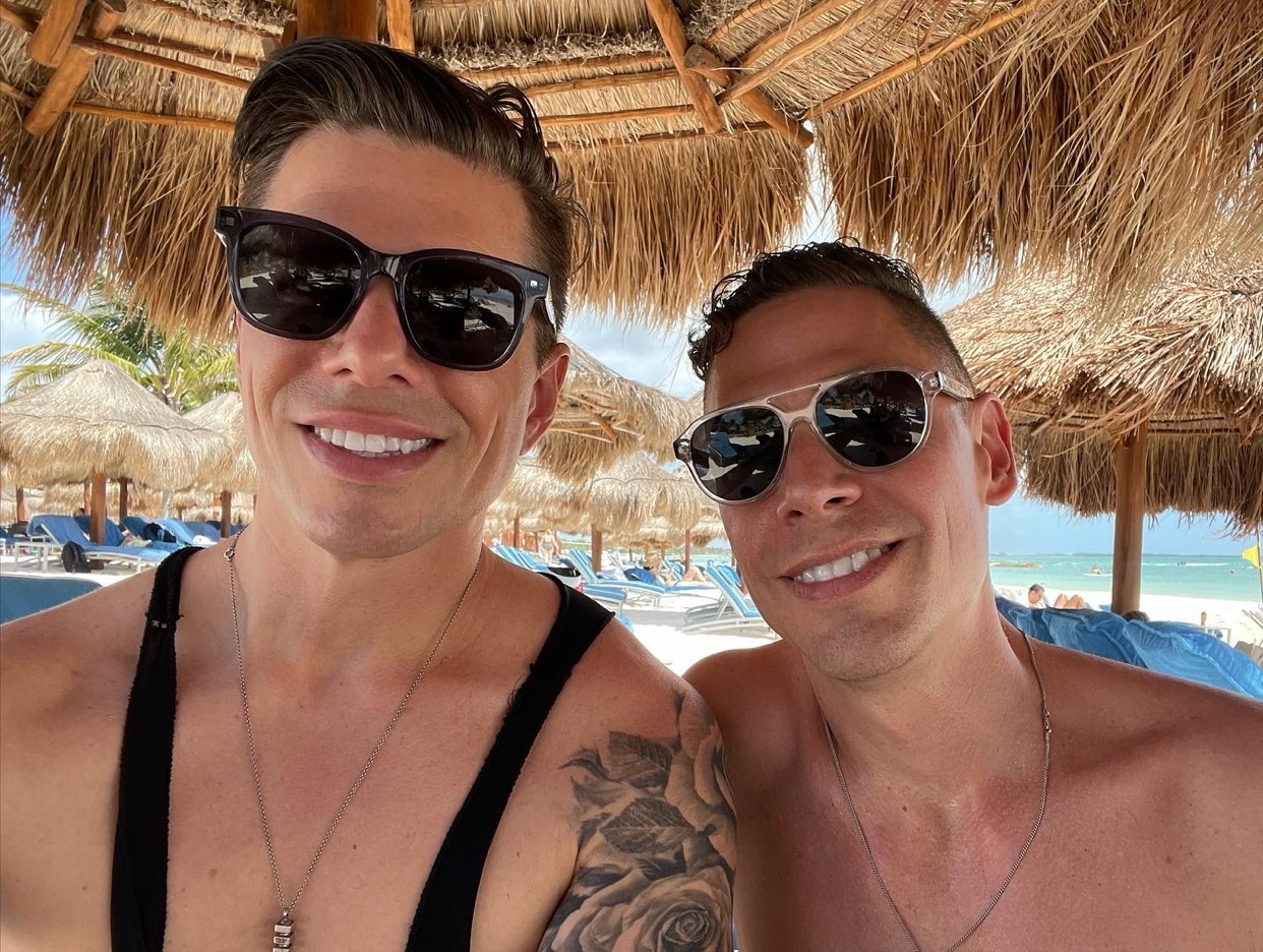 A close-up selfie of two men smiling at the camera, wearing sunglasses and standing on a beach with thatched umbrellas in the background.