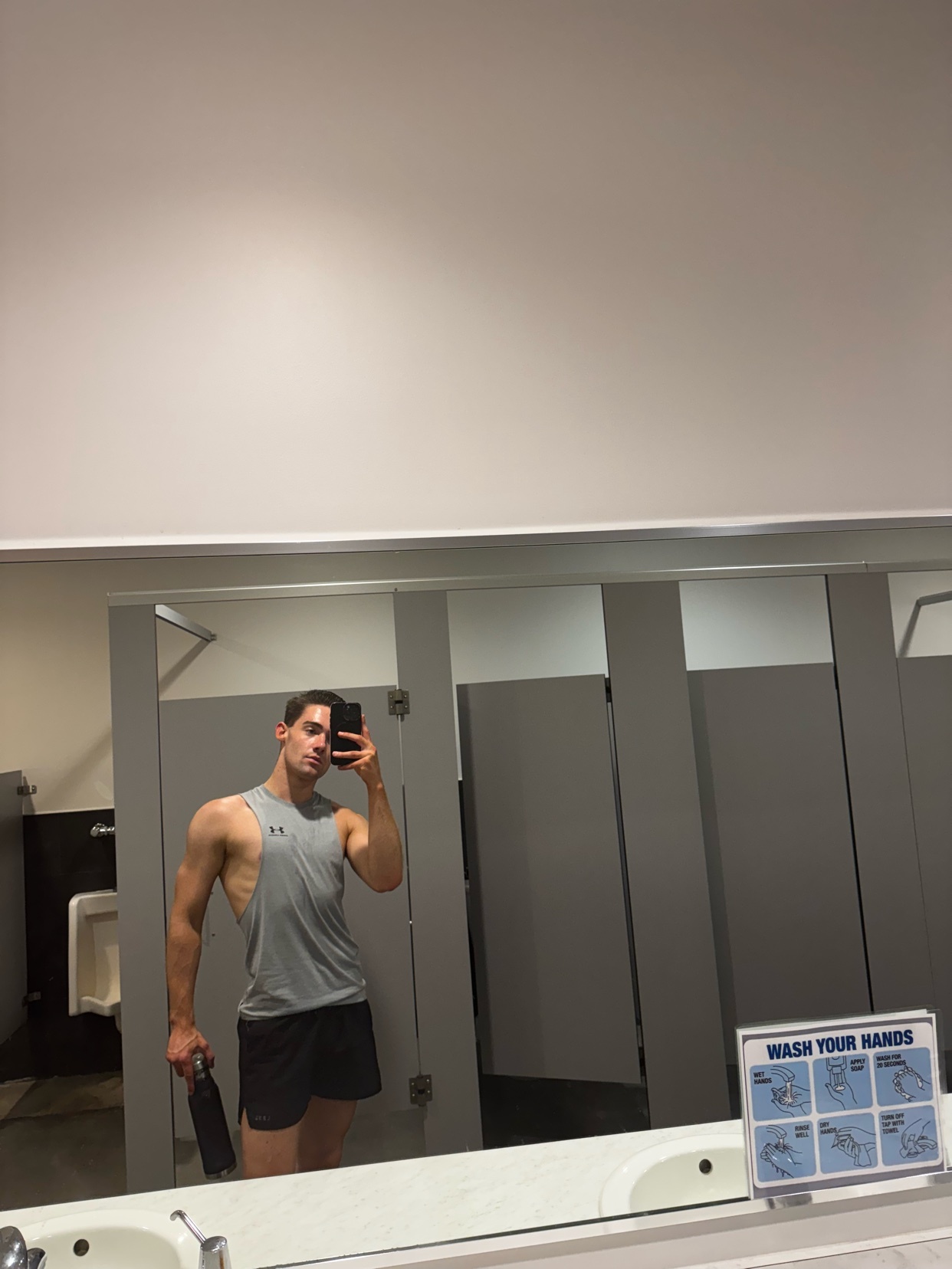 A man takes a mirror selfie in a gym restroom, holding a water bottle and wearing a tank top and shorts.