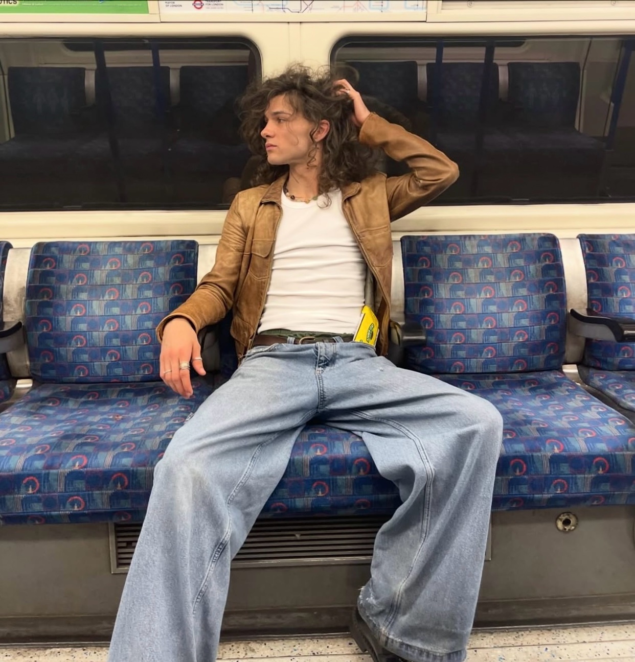 A man with long curly hair poses on a train, wearing a leather jacket and wide-leg jeans. The photo captures a stylish and modern aesthetic.
