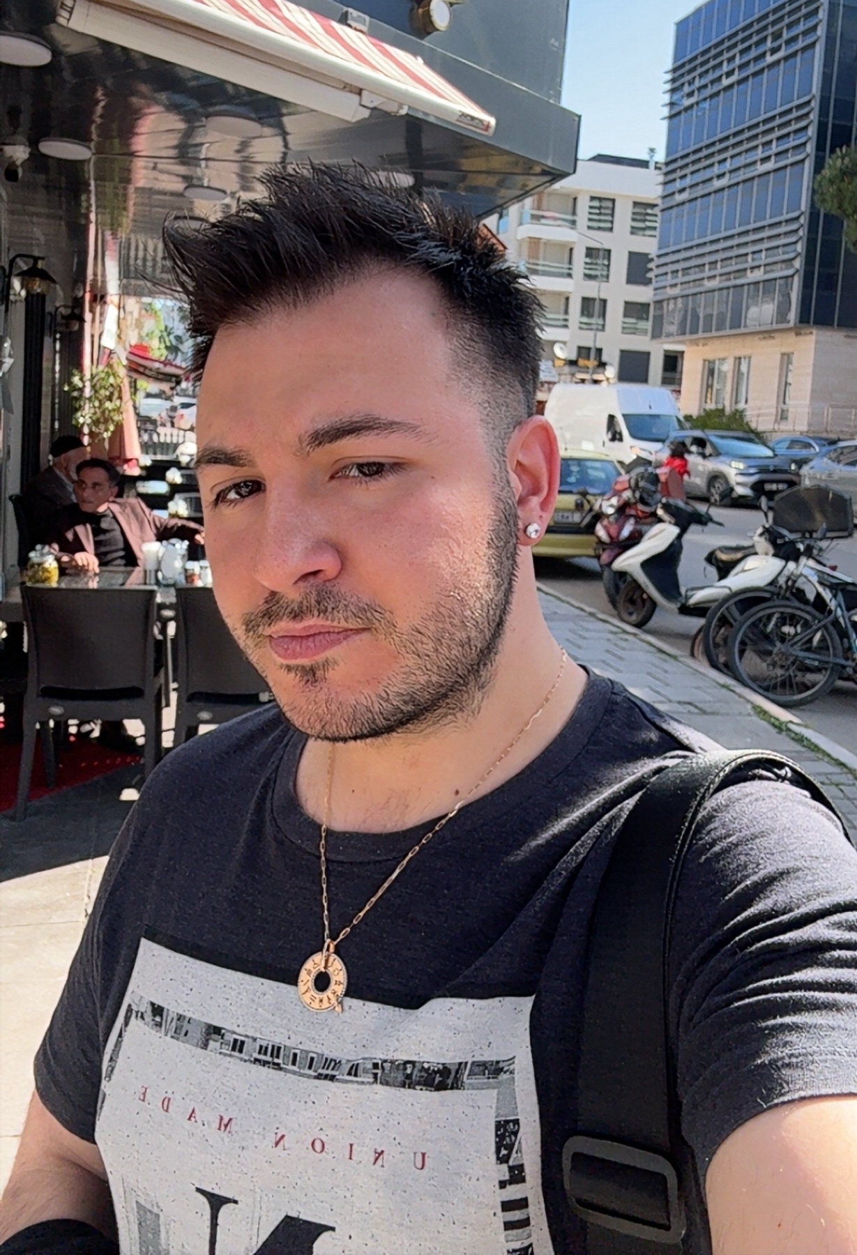 A man with short, dark hair and stubble takes a selfie outdoors on a sunny day. He is wearing a dark t-shirt with a graphic print and a gold necklace.
