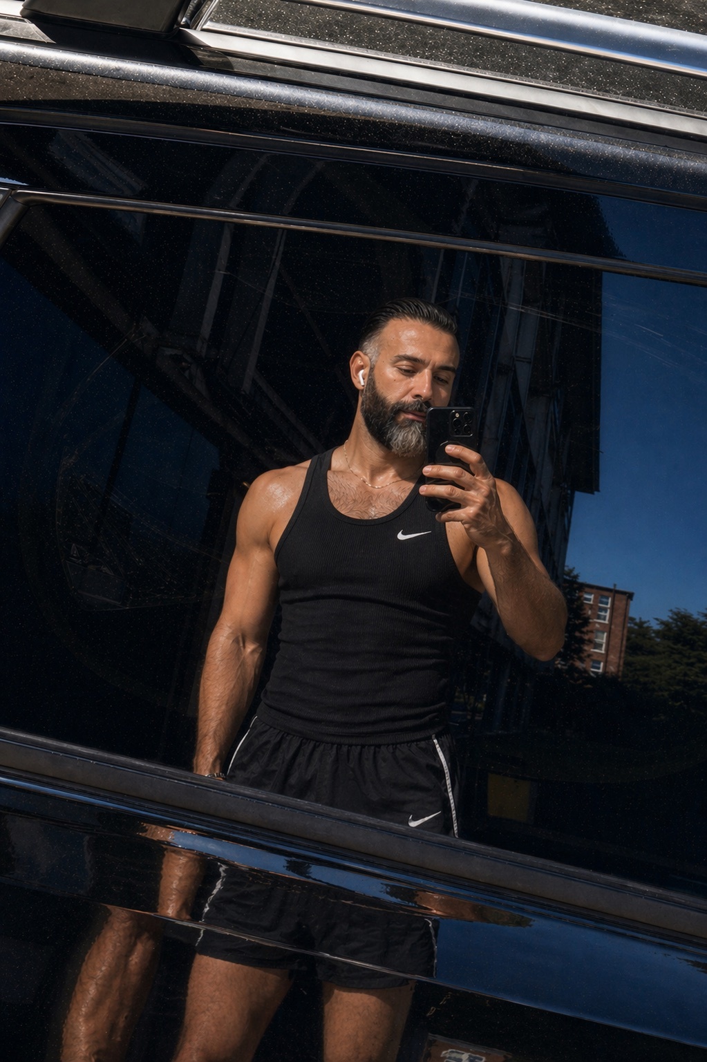 A man with a beard takes a selfie in the reflection of a car window. He is wearing a black tank top and shorts, and appears to be at a gym or workout location.