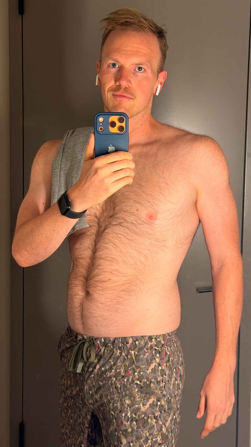 A man with a mustache takes a selfie in what appears to be a gym locker room. He is shirtless and wearing patterned shorts, with wireless earbuds in his ears.