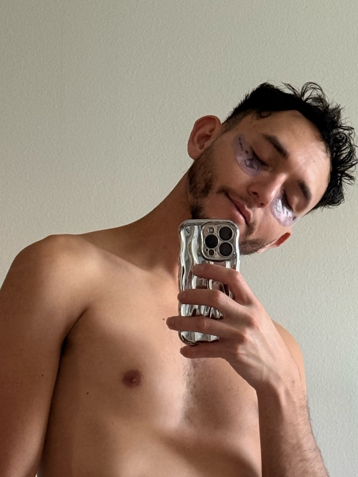 A man takes a selfie, shirtless, with eye patches on. The photo has a modern aesthetic.