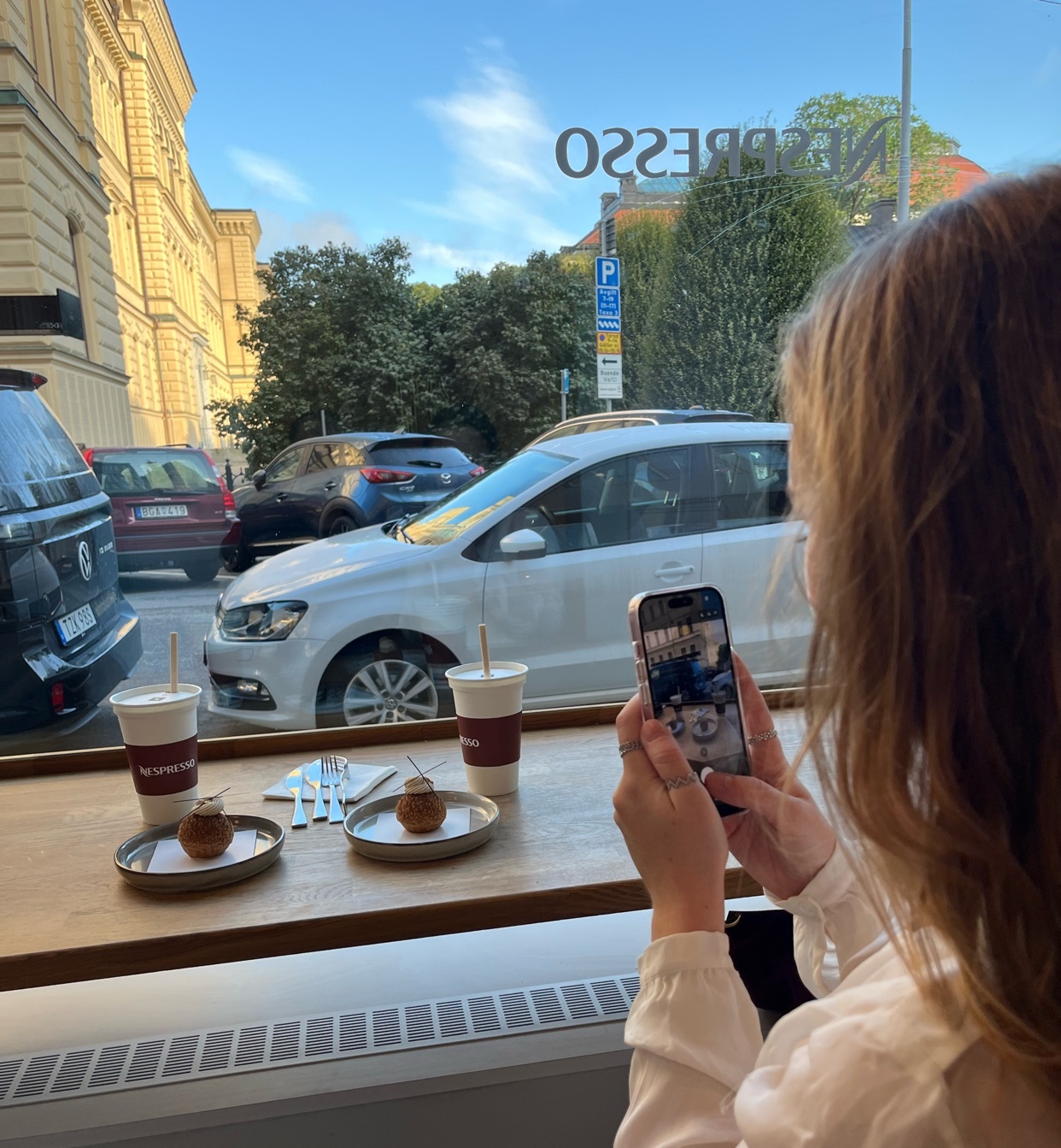 A woman in a white shirt is taking a photo of two Nespresso pumpkin spice iced lattes and two choux pastries on a table in front of a window. The pastries are small and round, topped with what looks like whipped cream and a sprinkle of cinnamon. The
