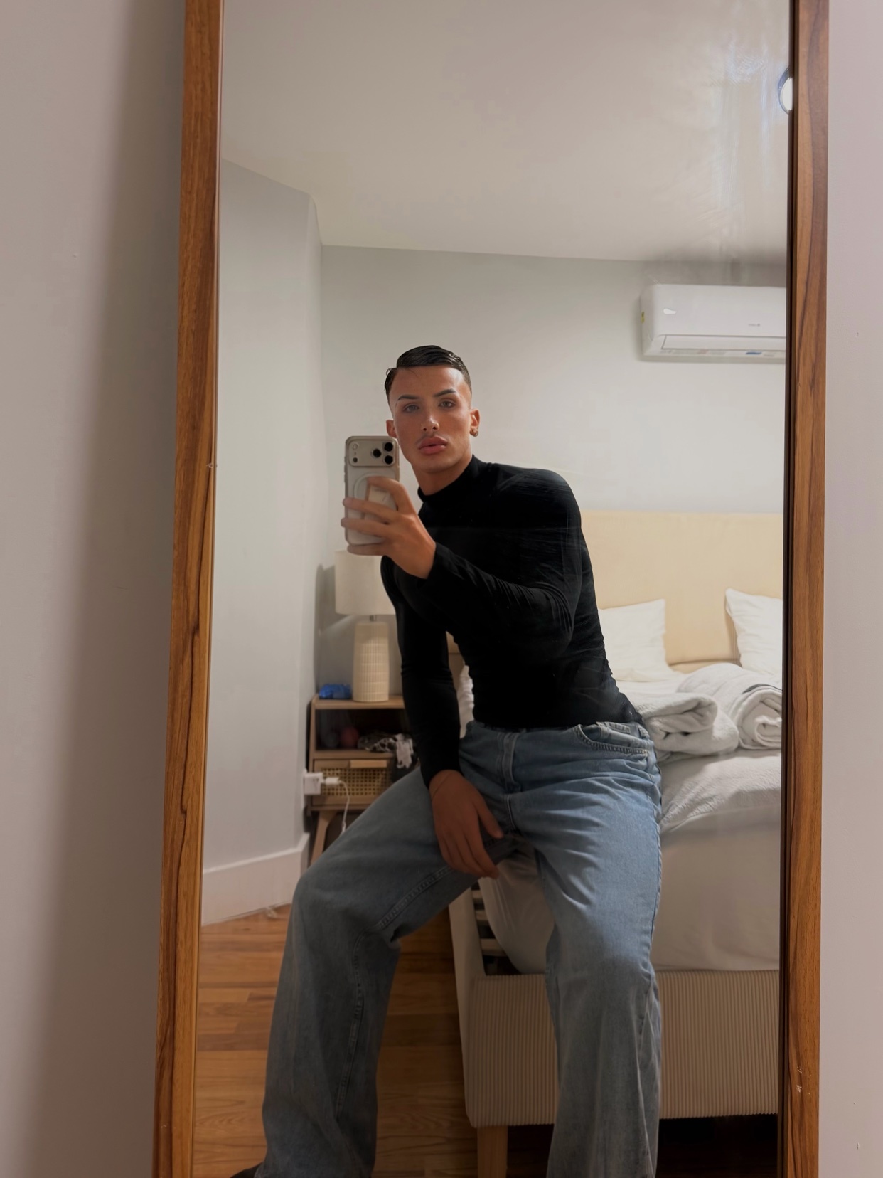 A man takes a mirror selfie in a bedroom, posing in a black turtleneck and baggy jeans.