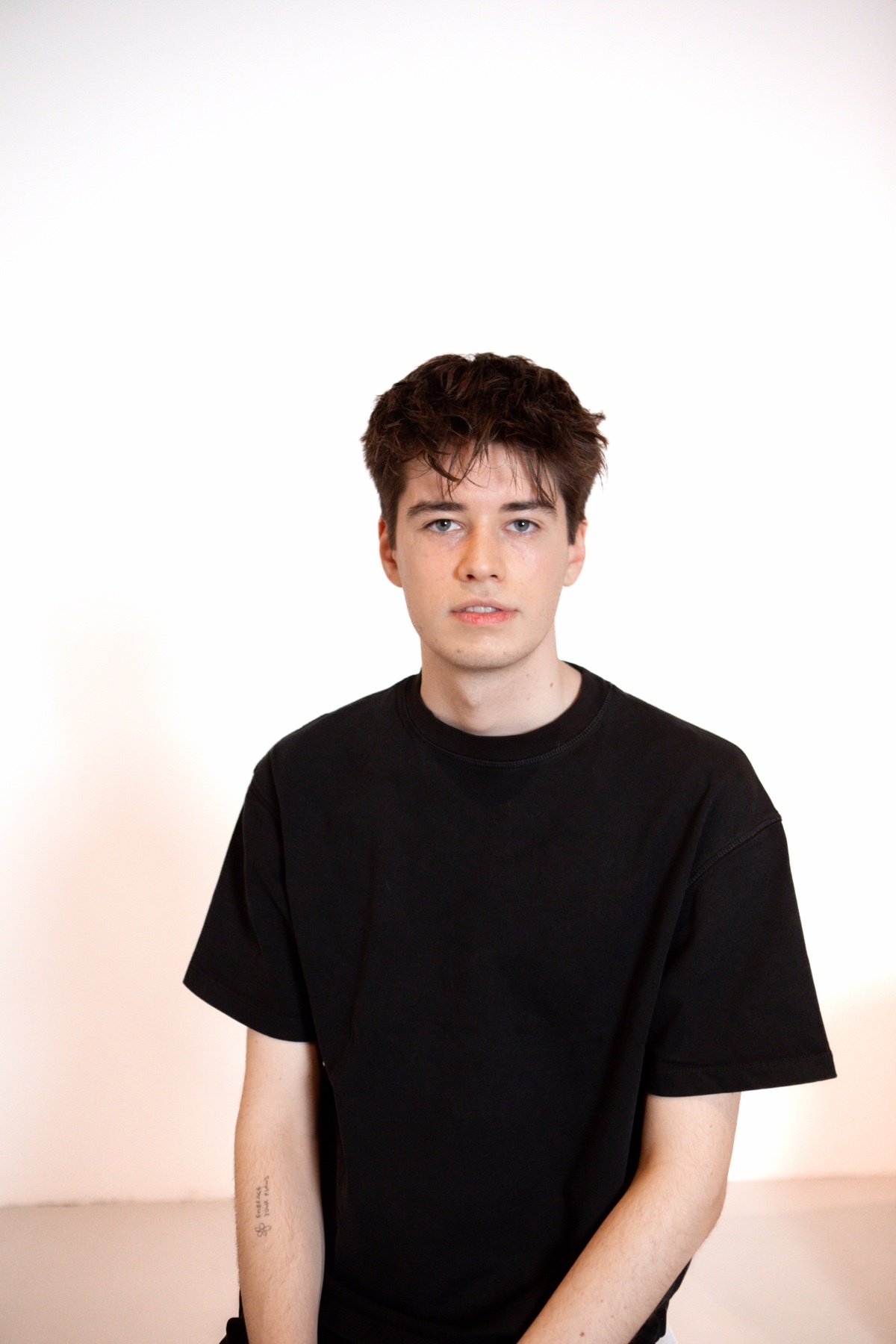 A man in a black t-shirt poses for a studio portrait. He has dark hair and a small tattoo on his arm.