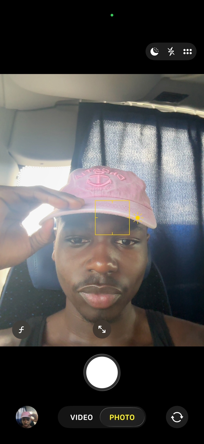 A young black man wearing a pink baseball cap and a black tank top takes a selfie. He is adjusting his cap with his left hand and looking directly at the camera.