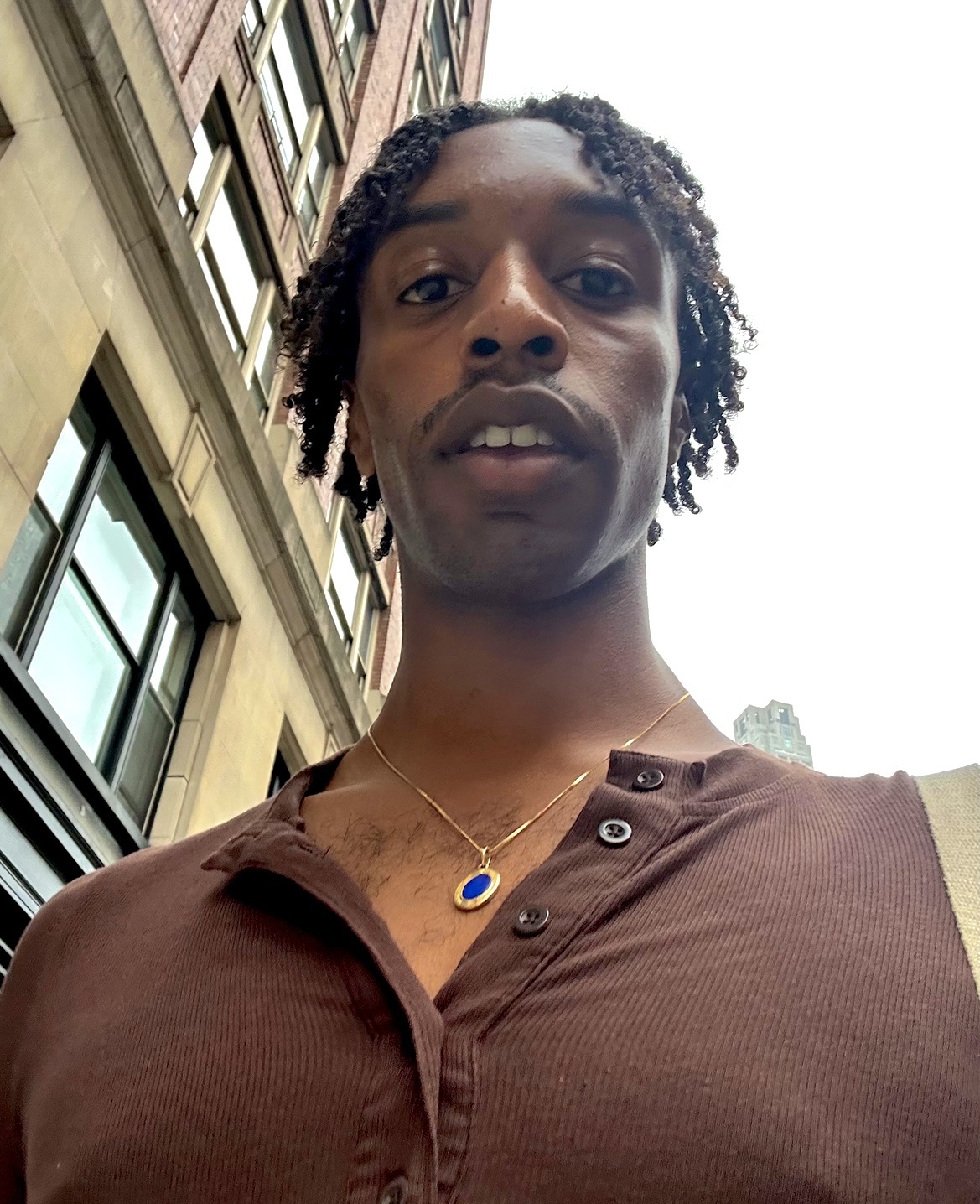 A man takes a selfie in front of a building. He is wearing a brown shirt and a necklace.