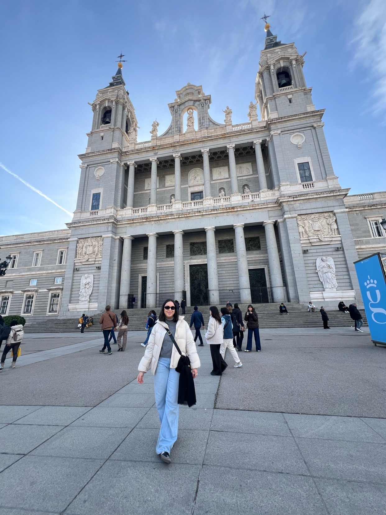 A woman poses in front of a large cathedral in Madrid, Spain. The photo captures a sunny day with a clear blue sky.