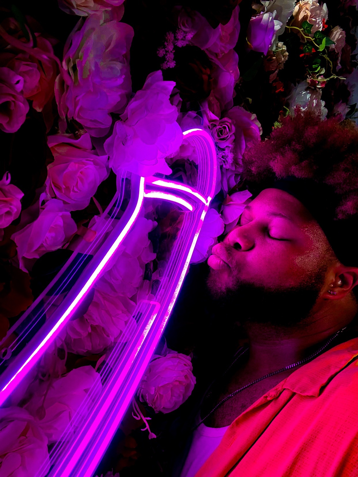 A man is kissing a neon light in front of a wall of flowers. The image is lit with pink and purple hues.