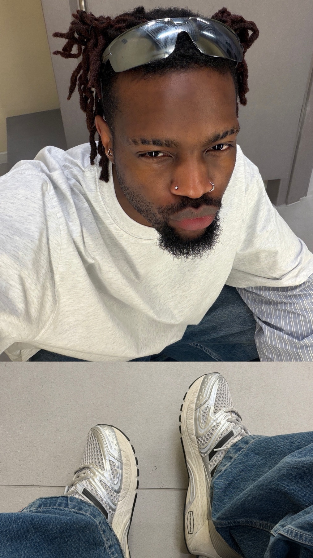A man with dreadlocks and a nose ring takes a selfie in a bathroom, wearing sunglasses on his head and a light grey t-shirt with blue jeans. The bottom half of the image shows his feet in silver sneakers.