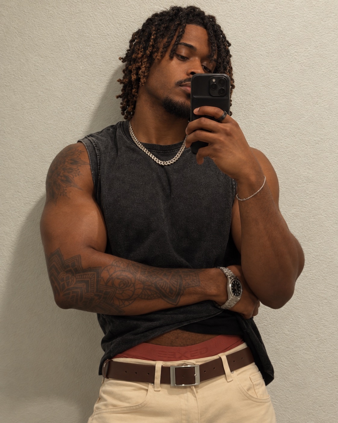 A man with dreadlocks and a beard takes a selfie in a sleeveless shirt and tan pants. He has tattoos on his arms and is wearing a watch and a silver chain.