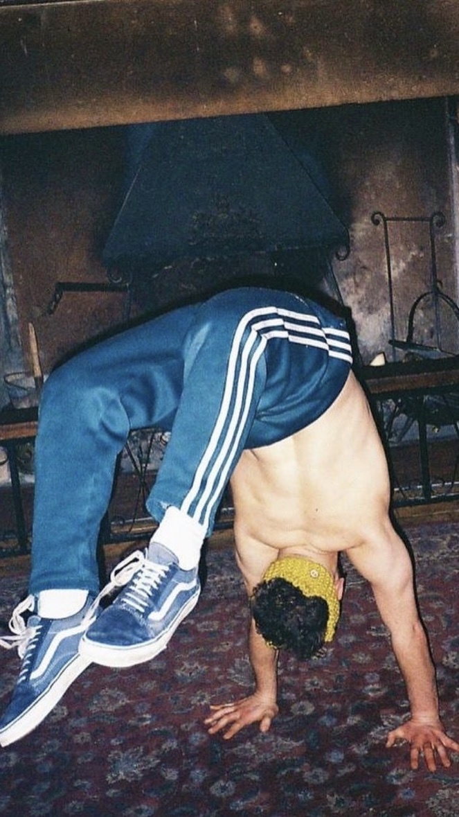 A shirtless man in athletic pants and Vans shoes performs a backbend on a patterned rug in front of a fireplace. He wears a yellow beanie and his arms are supporting his weight as he arches his back.