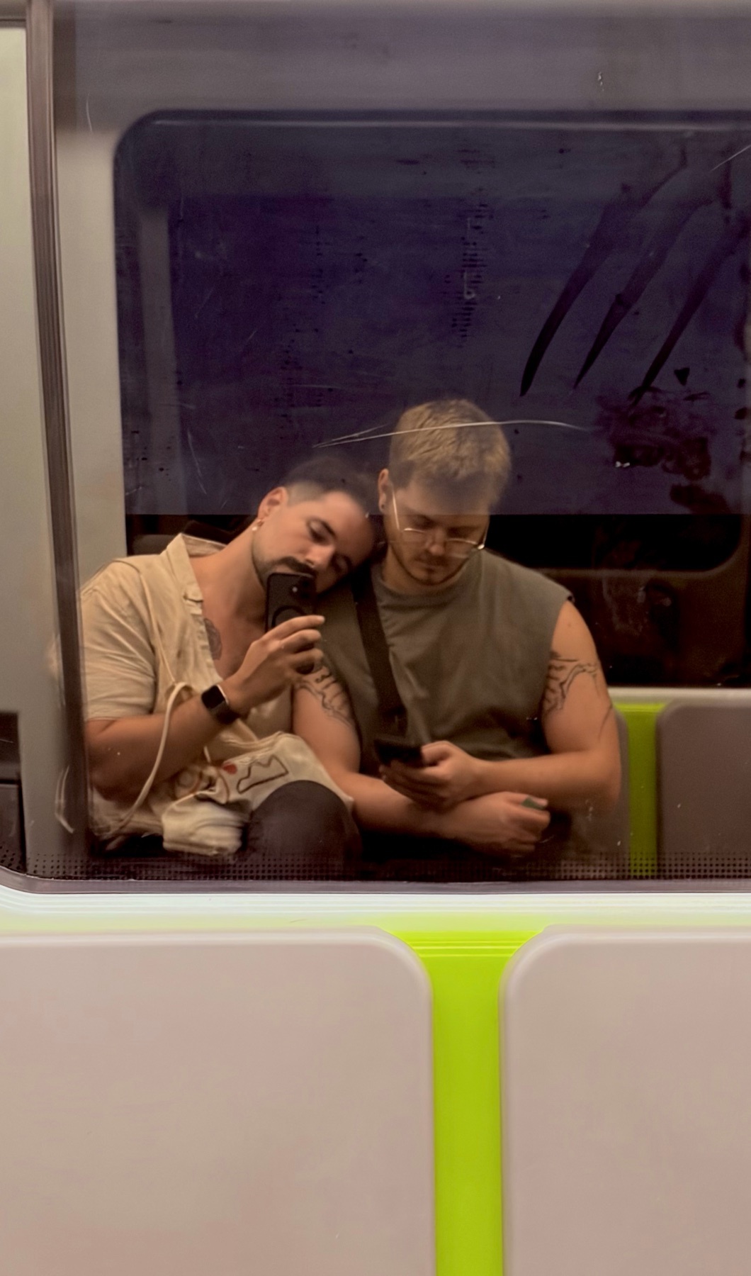 A couple is reflected in a train window, with one man resting his head on the other's shoulder while holding a phone. The other man is looking down at his phone.