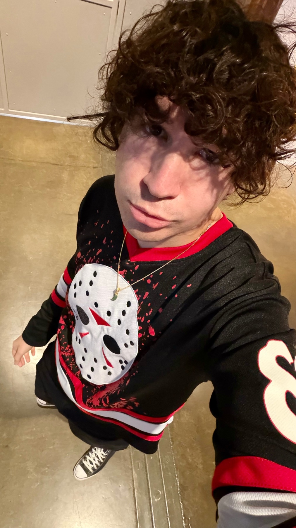 A young man with curly brown hair takes a selfie while wearing a black hockey jersey with a white Jason Voorhees mask graphic on it.