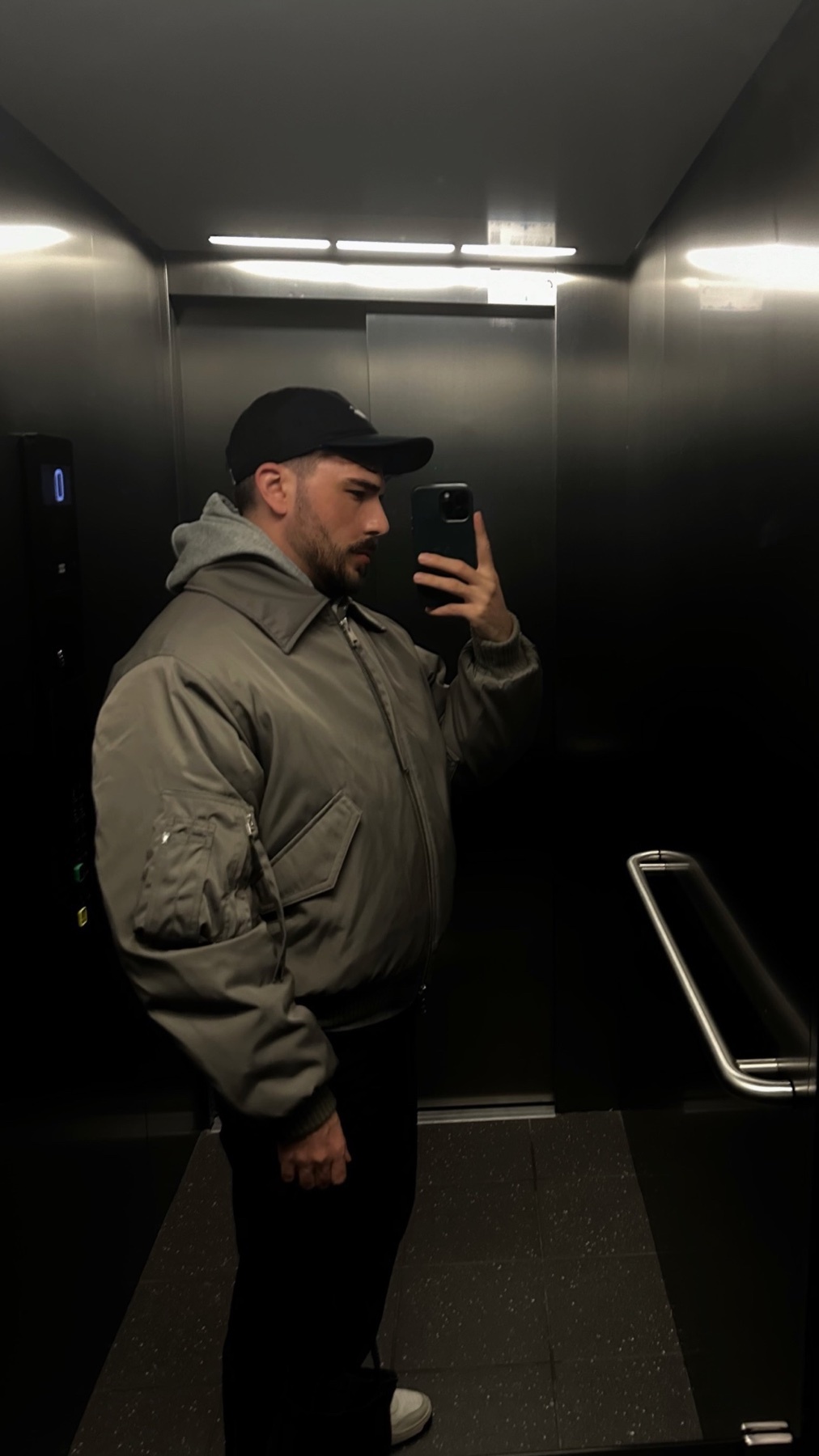A man takes a mirror selfie in an elevator. He is wearing a jacket and a cap.