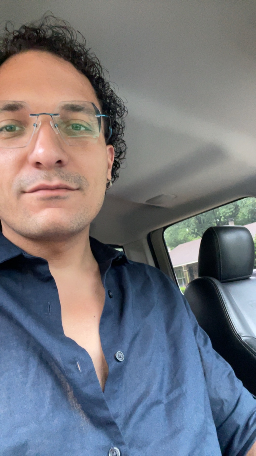 A man with curly dark hair and glasses takes a selfie in a car. He is wearing a dark blue button-up shirt.