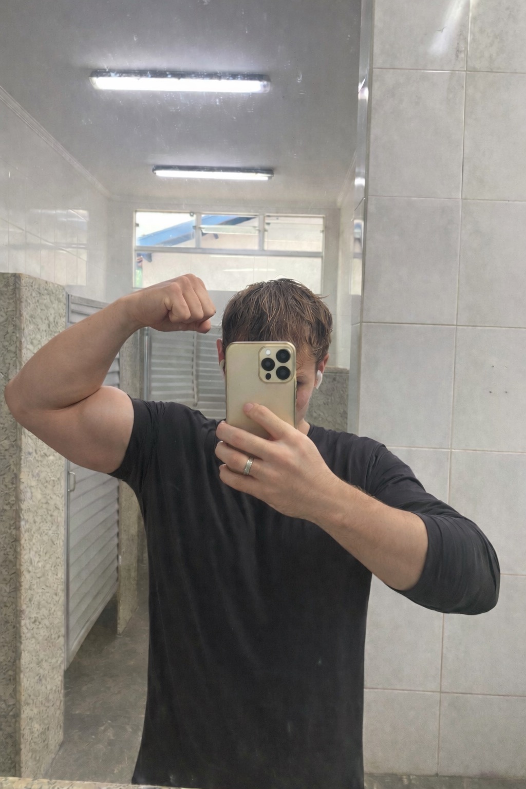 A man flexes his bicep in a mirror selfie taken in a gym bathroom. He is wearing a black t-shirt and has a ring on his left hand.
