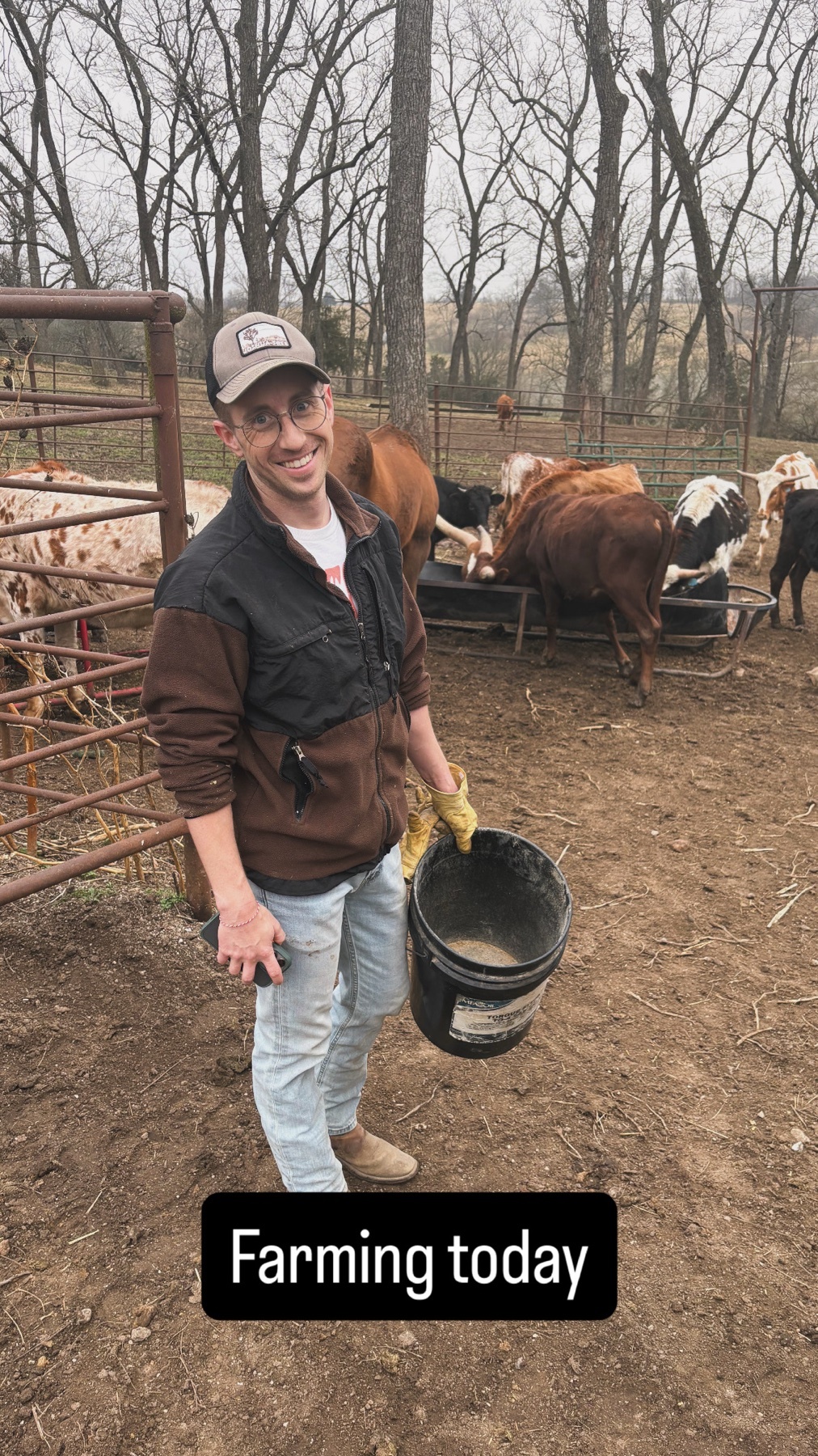 A man in a cowboy hat and work clothes smiles at the camera while holding a bucket of feed. He is standing in a muddy enclosure with cattle in the background.