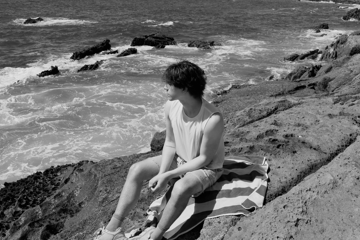A young man sits on a rocky coastline, looking out at the ocean. He is wearing a tank top and shorts, with a striped towel beneath him.