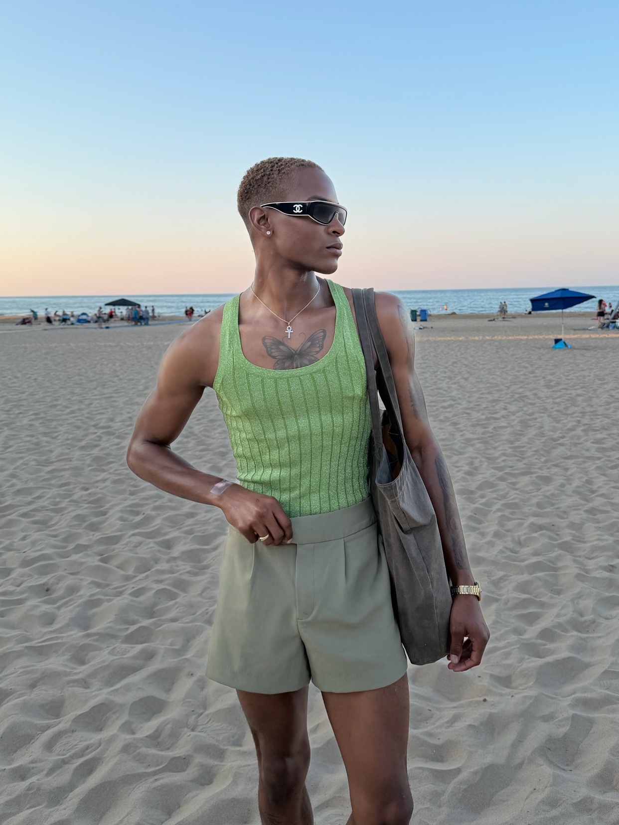 A man poses on a sandy beach wearing a lime green ribbed tank top and olive green shorts. He accessorizes with sunglasses, a necklace, a ring, and a watch, carrying a tote bag over his shoulder.