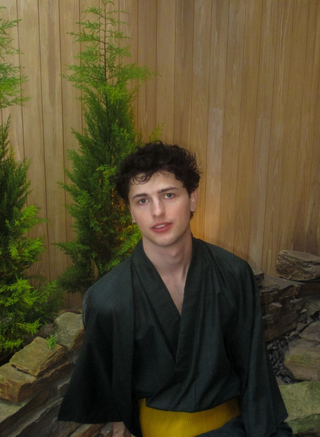 A young man with dark, curly hair poses in a dark green yukata with a yellow sash. He is indoors with a wooden wall and some greenery behind him.