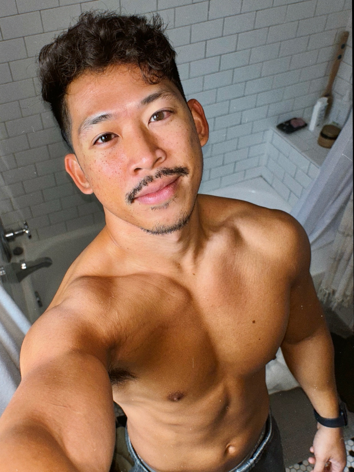 A man takes a shirtless selfie in a bathroom. He has a muscular physique and a slight smile.
