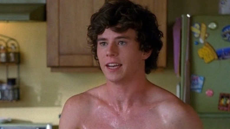 A young man with curly brown hair is shown from the chest up, looking off to the side with his mouth slightly open. He appears to be in a kitchen and is sweating.