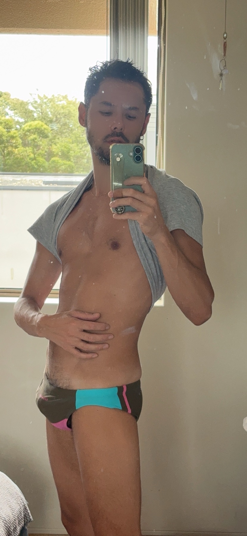 A man takes a mirror selfie wearing briefs and a grey t-shirt draped over his shoulders. He has stubble and dark hair.