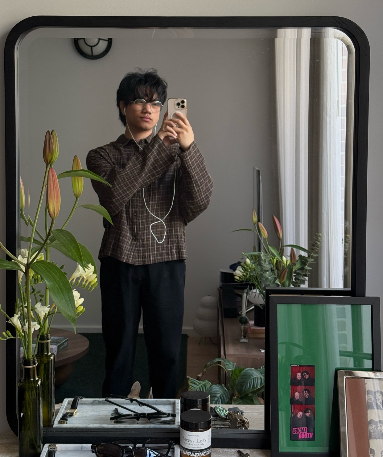 A young man takes a mirror selfie while wearing a plaid shirt and black pants. He has earbuds in and is wearing glasses.