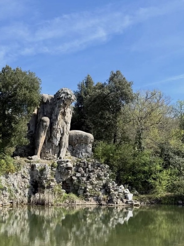 A large, weathered stone sculpture of a seated figure is nestled amongst rocks and lush green trees, with its reflection shimmering in the calm water below.