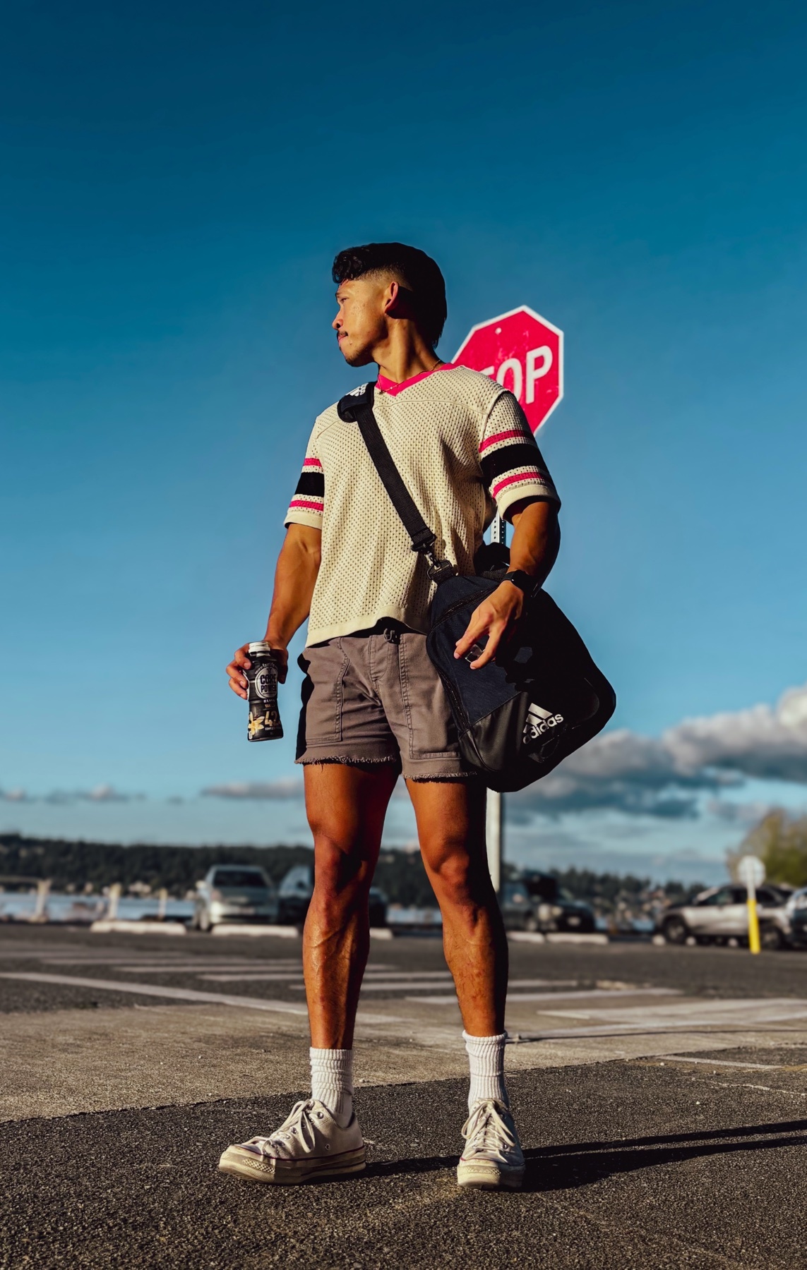 A man stands confidently, holding a drink and a gym bag, with a stop sign in the background. He is wearing shorts and a t-shirt, showcasing his muscular legs.