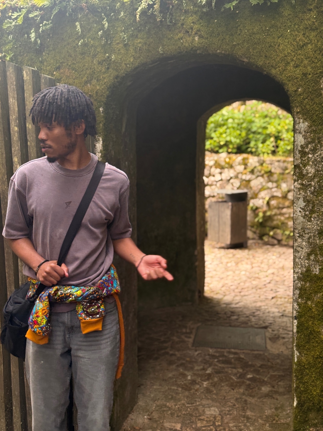 A man with dreadlocks stands in a stone archway covered in moss. He is wearing a purple t-shirt, grey jeans, and a colorful tie-dye sweatshirt tied around his waist. He is looking to his left and gesturing with his right hand.