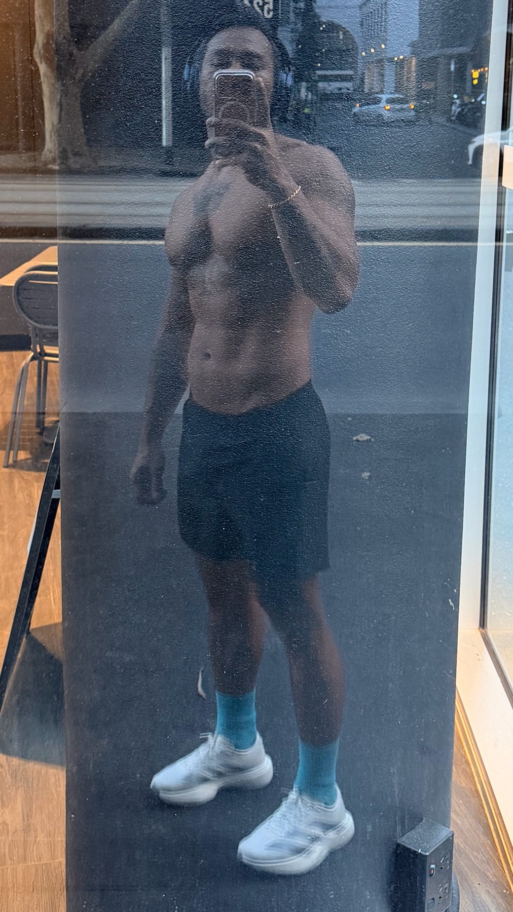 A shirtless man with headphones on takes a mirror selfie after an evening run. He is wearing black shorts and blue socks with white athletic shoes.