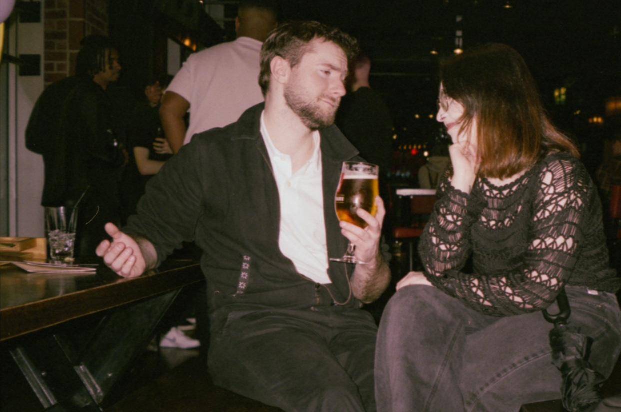 A man and a woman are sitting at a table, engaged in conversation. The man holds a glass of beer, and the background suggests a lively indoor setting.