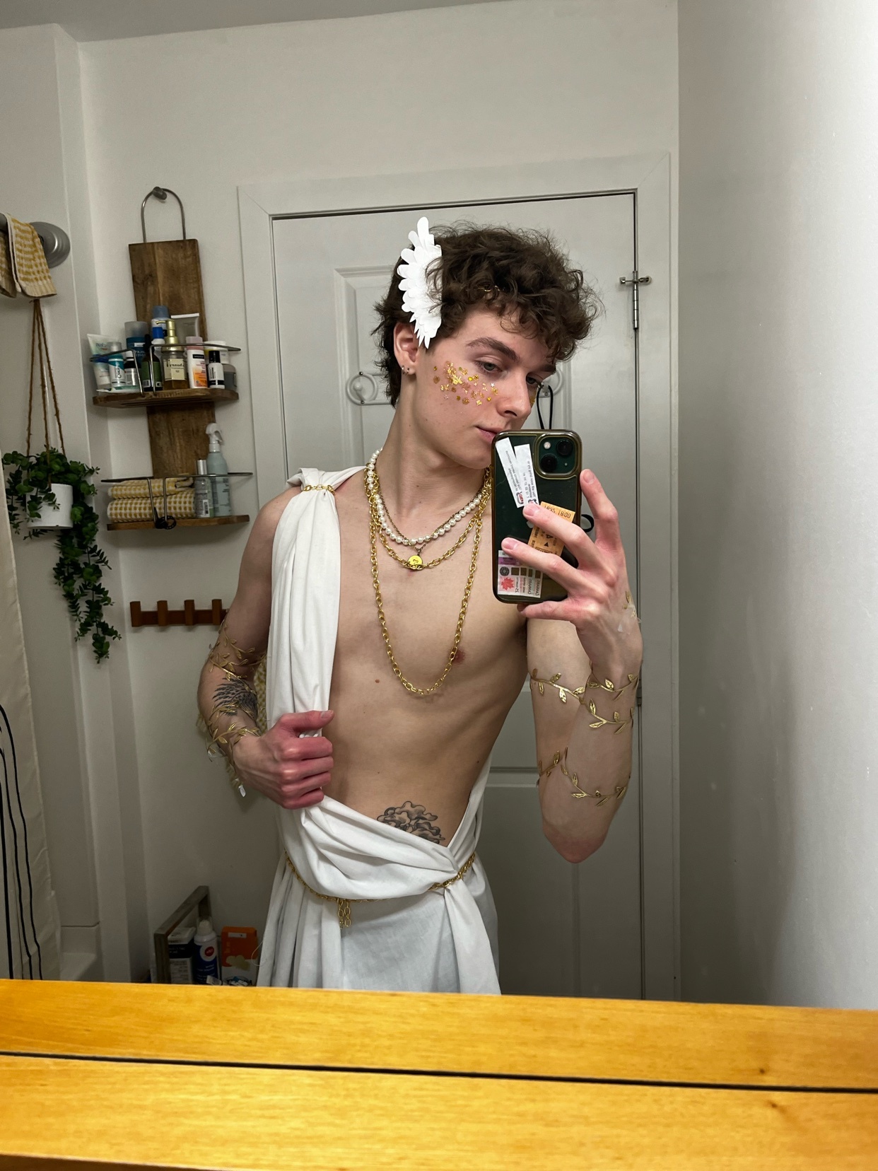 A young man in a white toga and gold jewelry poses for a selfie in a bathroom, dressed as a Greek god.