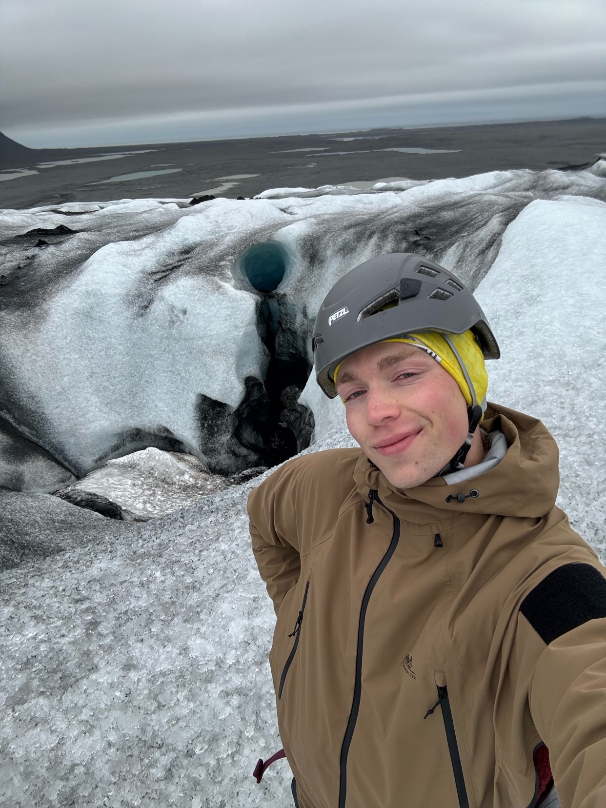 A man in a tan jacket and yellow beanie smiles at the camera while standing on a glacier. He is wearing a grey helmet and the background shows a vast icy landscape with a small blue pool of water.