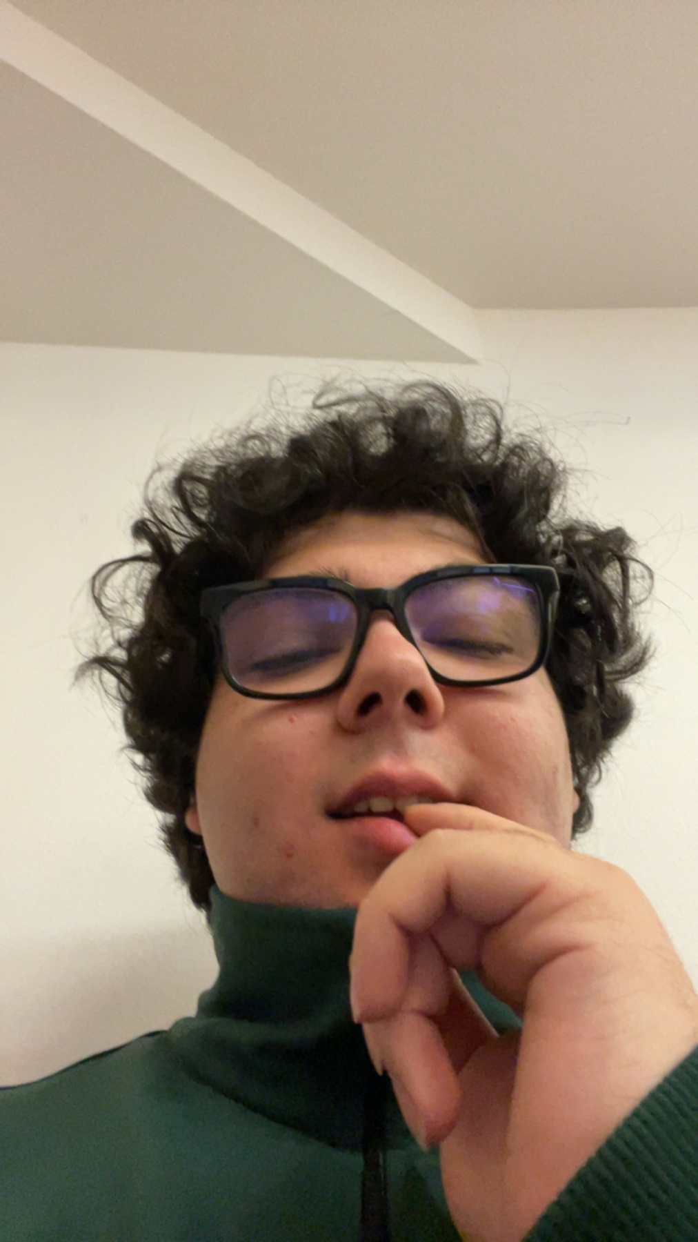 A selfie of a young man with curly black hair and glasses. He is wearing a green turtleneck.