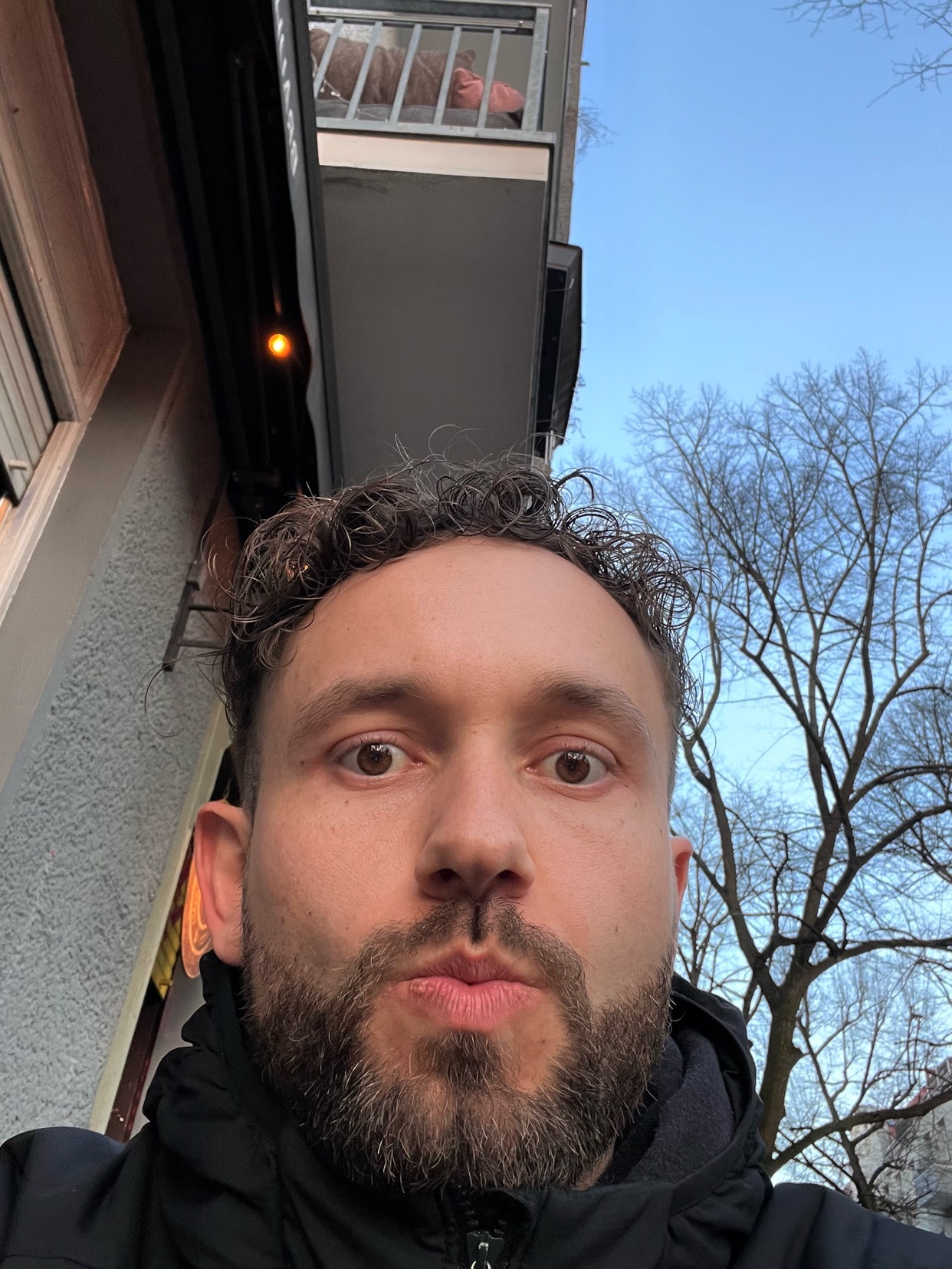 A selfie of a man with curly hair and a beard. The man is making a funny face.