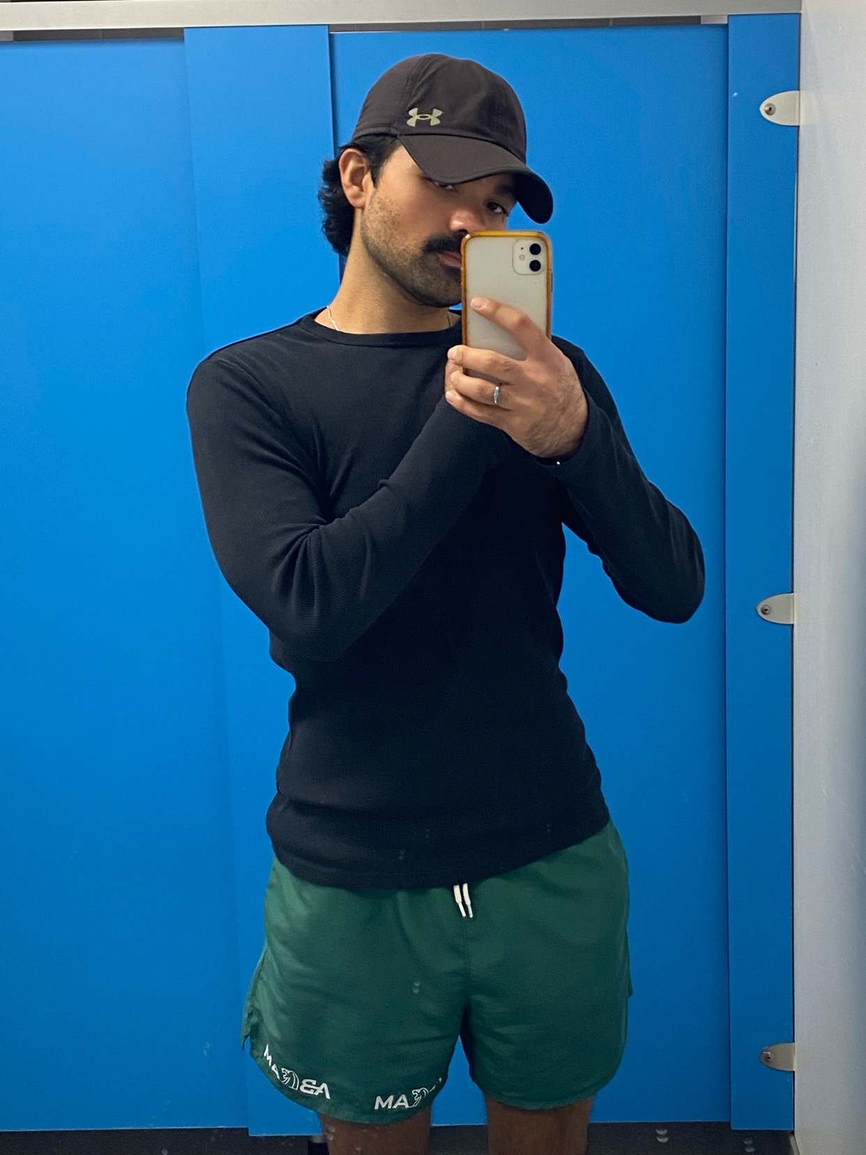 A man in a black long-sleeved shirt, green athletic shorts, and a black baseball cap takes a mirror selfie in what appears to be a locker room. He has a mustache and a beard.