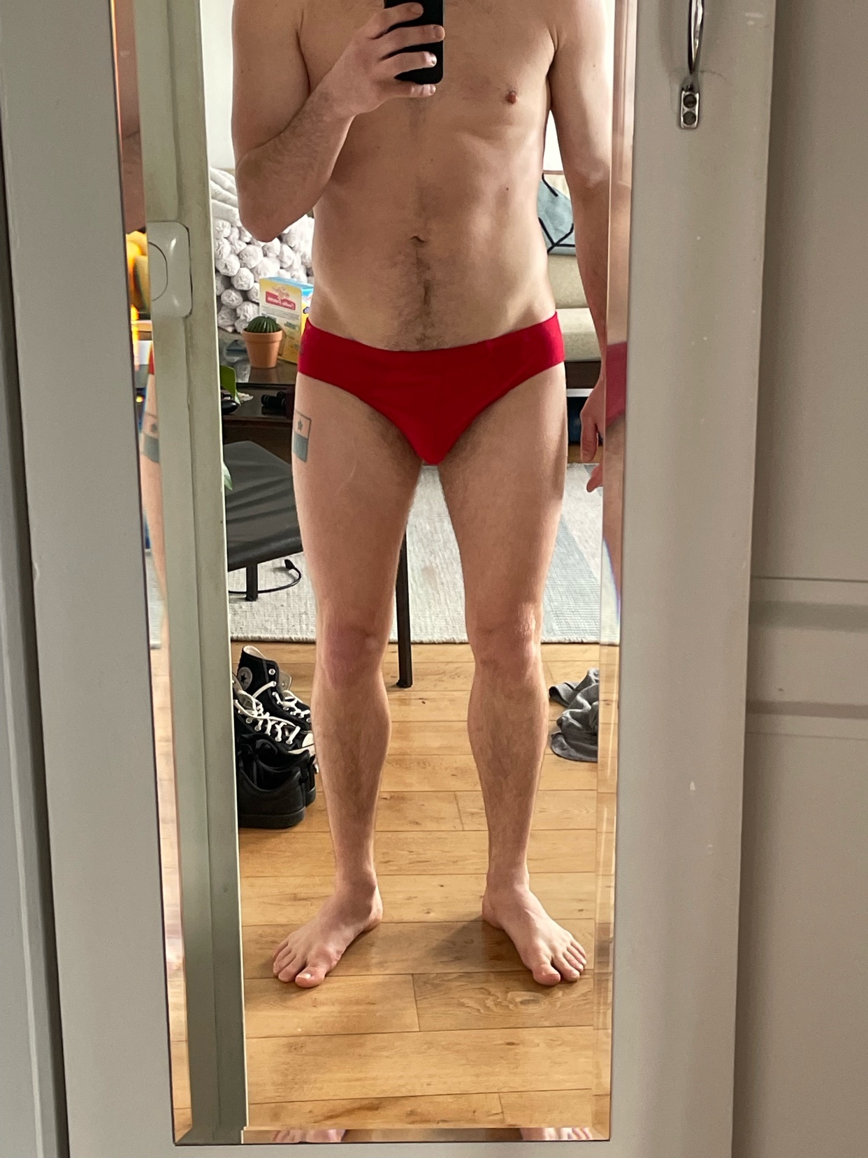 A man in red swimwear poses for a mirror selfie. He is shirtless, showing his torso and legs. A tattoo is visible on his left thigh.
