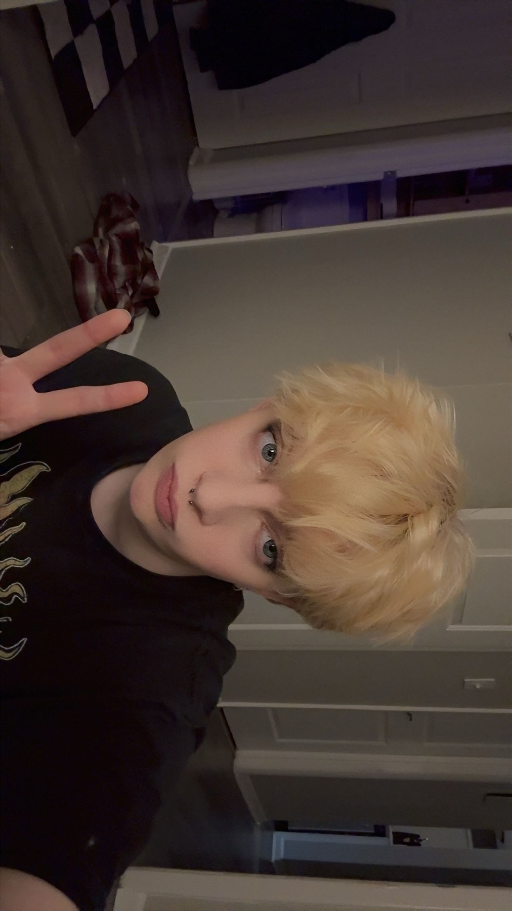 A person with short, blonde hair and blue eyes poses for a selfie, holding up a peace sign.